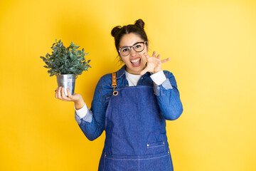 Young caucasian gardener woman holding a plant isolated on yellow background smiling funny doing...
