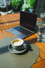 A cup of aromatic coffee on the background of a laptop on a summer terrace. Work in a cafe. Remote work online.