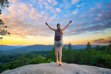 Sportive woman standing alone on hillside trail with raised up arms at sunset. Female hiker...