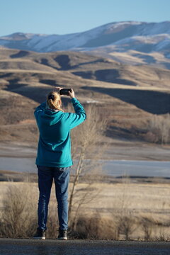 A girl in a hooded sweater and jeans takes pictures of the lake and mountains on the side of the road