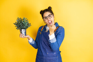 Young caucasian gardener woman holding a plant isolated on yellow background showing middle finger...