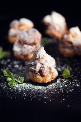 Profiteroles or cream puff with filling,  falling powder sugar topping. Berries, mint on black background. Fresh homemade Cream Puffs, cake, tasty French choux puff, ecler, dessert closeup. Vertical 