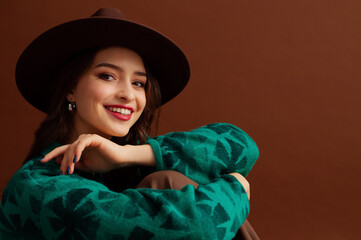 Fashionable happy smiling woman wearing hat, trendy silver hoop earrings, green sweater, posing on brown background. Close up studio fashion portrait. Copy, empty, blank space for text
