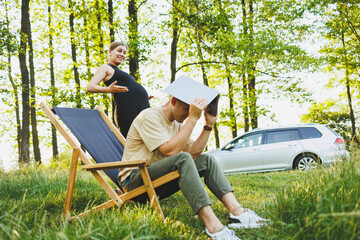 A happy couple relaxes in folding chairs near the car, a man uses a laptop for work or checking social networks, his wife treats him to a croissant. Pregnant woman with man in nature. - obrazy, fototapety, plakaty