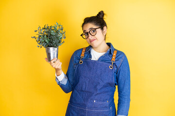 Young caucasian gardener woman holding a plant isolated on yellow background with angry face,...