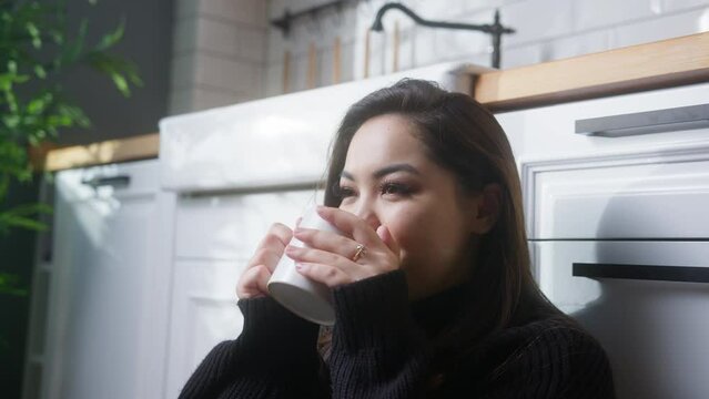 Relaxed young asian woman sitting on floor in kitchen, drinking morning coffee or hot tea, enjoying spending time in own house.