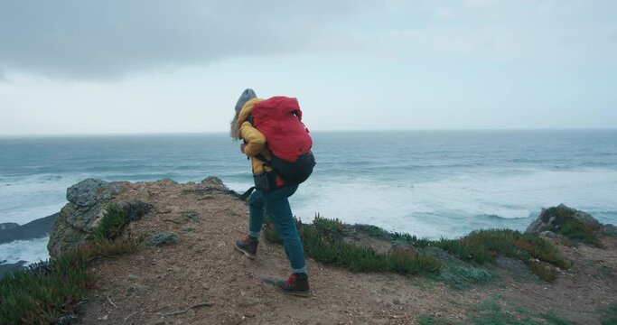 Inspiring female hiker walks uphill along the ocean, stands on the edge of a cliff, and looks into the horizon. Adventurer reach top of the mountain. Empowered female explorer.