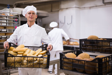 Baker in uniform holding crate with bread, industrial kitchen of bakery on background