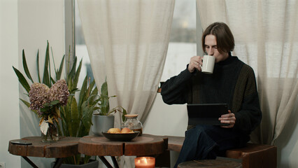 Relaxed caucasian man in modern kitchen interior enjoying tea and using tablet. Media. Young freelancer having a break from work at home.