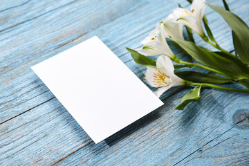 An elegant blank white card mockup, set on a blue wooden surface with white Alstroemeria flowers...