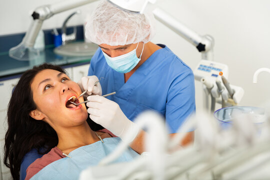 Man dentist in a protective mask working at the clinic examines a female patient with the help of working tools sitting in a ..dental chair. Close-up image