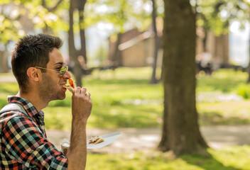 Handsome young man eating a slice of pizza outside on the street. Pizza time. Student having lunch break.  