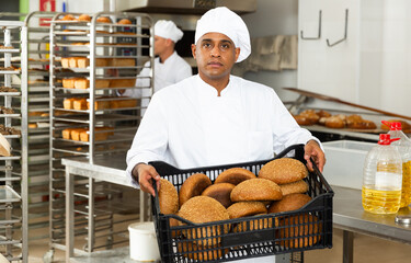 Focused young man working in small bakery, carrying fresh baked bread in crate