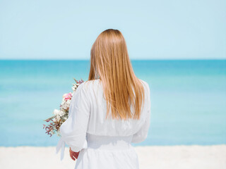 Fototapeta na wymiar Joyful Young Woman Holding a Bouquet of Flowers on a Beach with Blue Sky Background