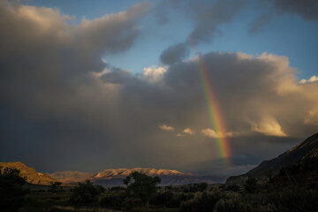 A striking rainbow emerges from dark storm clouds over a rugged landscape, casting a serene glow on the mountains at sunset.