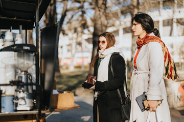 Two stylish women stand by a food truck, eagerly waiting to buy coffee in the warmth of the sun. A...