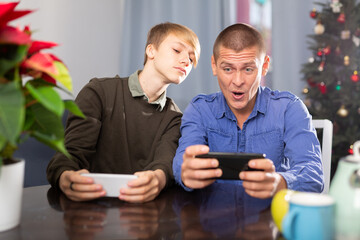 Happy father and son together playing on their smartphones while sitting at the table at home during the celebration of christmas