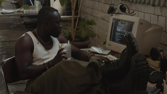 Young African American man sitting with legs on desk, eating canned food from foil pouch and running code on old computer while living in post apocalypse shelter