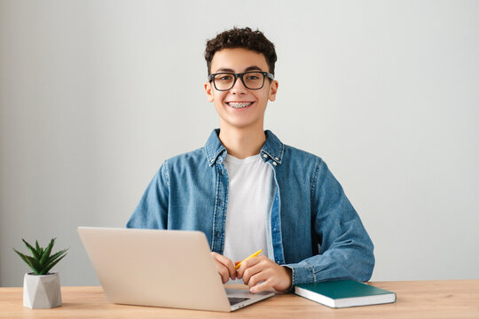 Smiling curly haired boy, teenager with dental braces on teeth using laptop computer studying