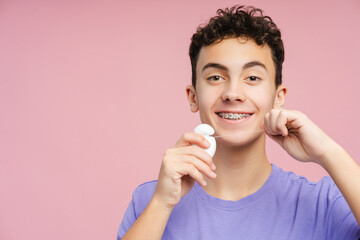 Smiling teenager with braces, carefully flossing teeth, isolated on pink background