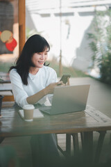 Business freelance concept, A happy woman engages with her smartphone while working on a laptop in a cozy, sunlit workspace with a coffee cup nearby.