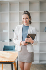 Business management concept, A smiling young professional woman stands in a modern office, holding a tablet, ready for a productive workday.