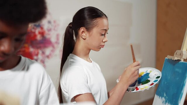 Multicultural student paint the canvas together in art lesson with colorful stained wall. Closeup of african boy and caucasian girl drawing picture or doing creative activity together. Edification