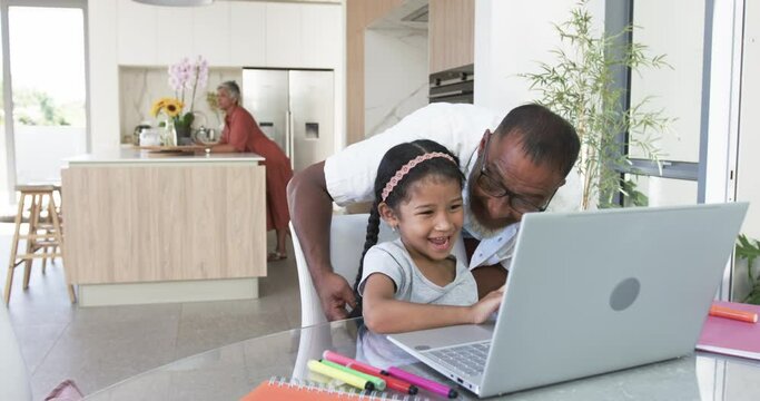 Biracial grandfather helps a young biracial granddaughter with a laptop in a modern kitchen