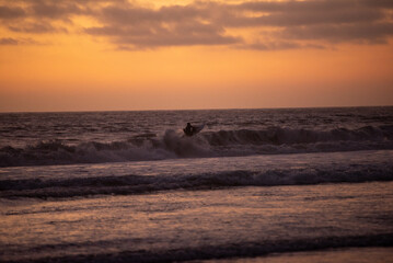 surfer while waves crash in pacific ocean with beautiful orange sunset with dark clouds
