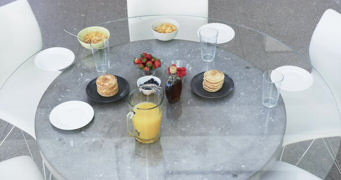 A breakfast spread is laid out on a glass table with pancakes, fruits, and juice