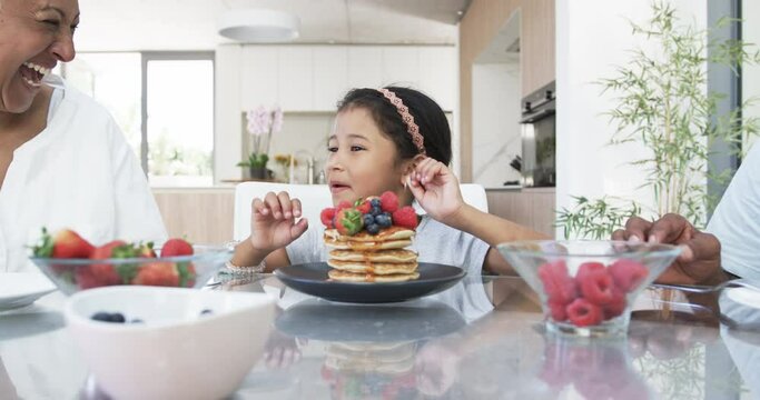A biracial child admires a stack of pancakes topped with berries at a kitchen table