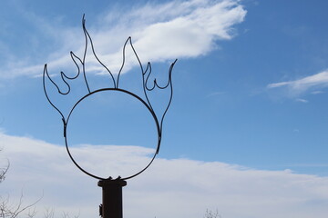Aro de metal con forma de flamas con cielo azul y nubes.