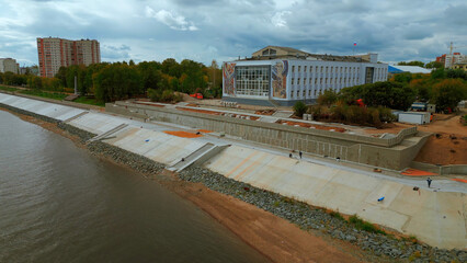 Aerial view of city embankment with construction site. Clip. Unfinished area and calm river with...