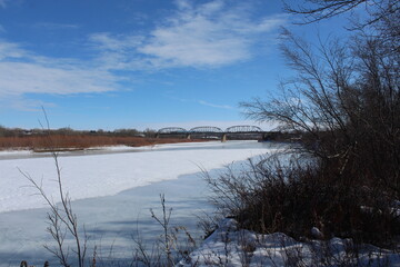 Paisaje con rio congelado y nevado con puentes al fondo y cielo azul.