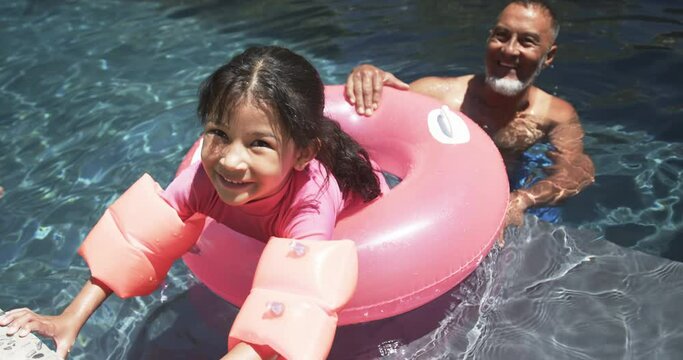A biracial girl with arm floaties enjoys pool time with her grandfather