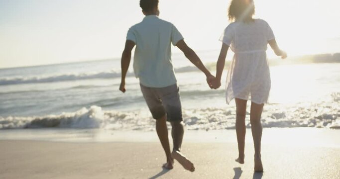 Biracial couple holding hands, walking on a sunlit beach