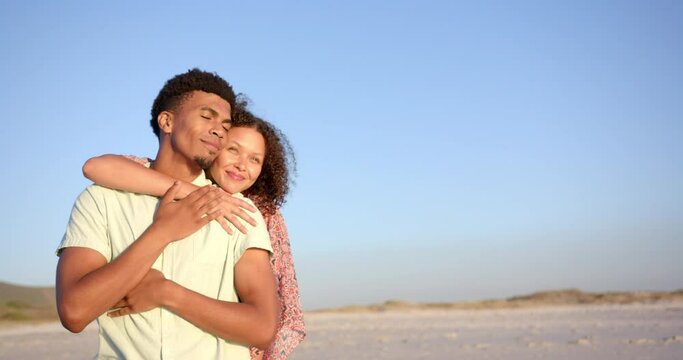 Biracial couple embraces warmly on a sunlit beach, eyes closed in contentment, with copy space