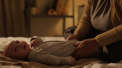 Medium close-up shot of happy African American mom sitting on bed at home with newborn baby son or daughter, stroking and massaging legs, then bending over, kissing on face and speaking lovingly - Powered by Adobe