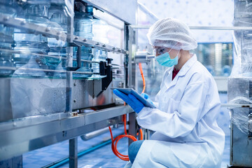 Female worker using tablet checking quality drinking water before process of filling water into plastic bottles to bring out to consumers. Water bottles on production line of factory.