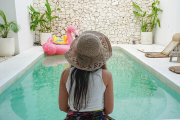 Back portrait of young Asian woman wearing white top with light cream colored hat sitting by swimming pool in a villa, relaxing and enjoying view for summer vacation - Powered by Adobe