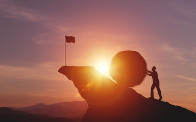 Silhouette of businessman pushing a large rock up hill with flag showing finish line on sunset...