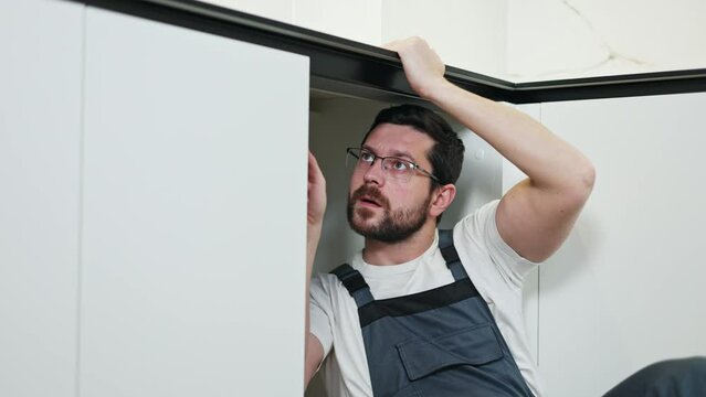 Focused workman holding countertop with hand while installing water sewer riser behind kitchen cabinet. Professional guy in spectacle finishing sanitary checking process and making apartment livable.
