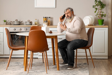 Portrait of sad senior man talking by phone in kitchen