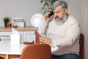 Portrait of sad senior man with empty wallet talking by phone in kitchen