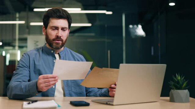Male director opening envelop and reading letter while sitting at desk with computer in office. Puzzled man receiving bad news and looking at document while thinking about solution of problem.