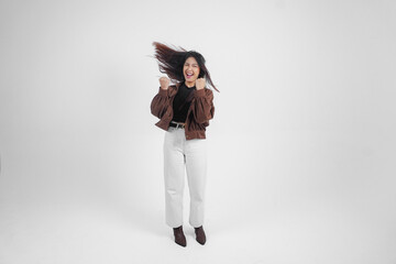 Active Asian girl is jumping and raising hands up wear brown jacket, isolated white color background