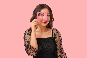 Beautiful young Asian woman holding paper sheet with hole in shape of heart on pink background. Valentine's Day celebration