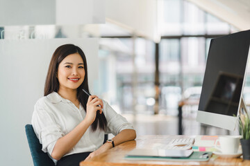 Obraz premium Smiling asian businesswoman in a casual white blouse sits confidently at her desk with a computer in a bright, modern office.