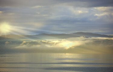 Fototapeta premium Sea, sunset and cloud on mountains in morning, reflection and island with landscape in nature. Blue sky, clouds or sunlight on water on beach, calm or sunrays on sustainable environment or seascape