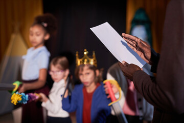 Close up of drama teacher holding play script with group of children on stage in background copy...